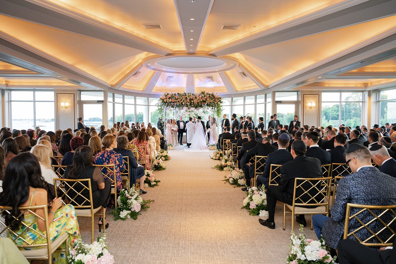 A wide view of the ceremony in full swing, aisle florals and gold chairs filling the glass ballroom.