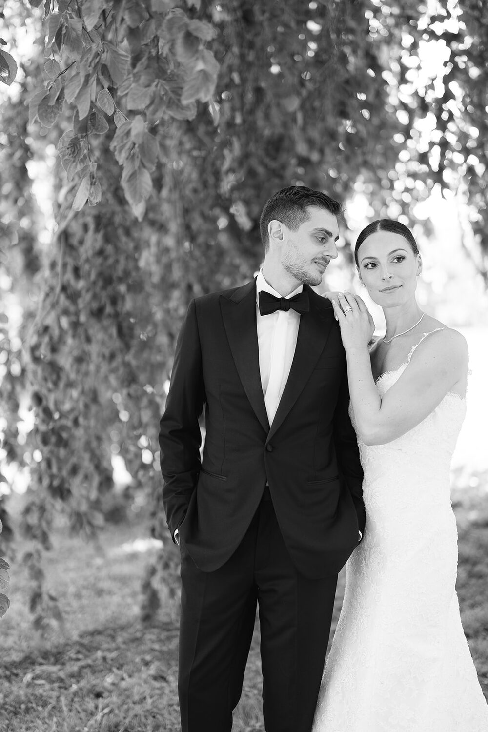 The bride rests her cheek on the groom's shoulder in the shade of a willow.