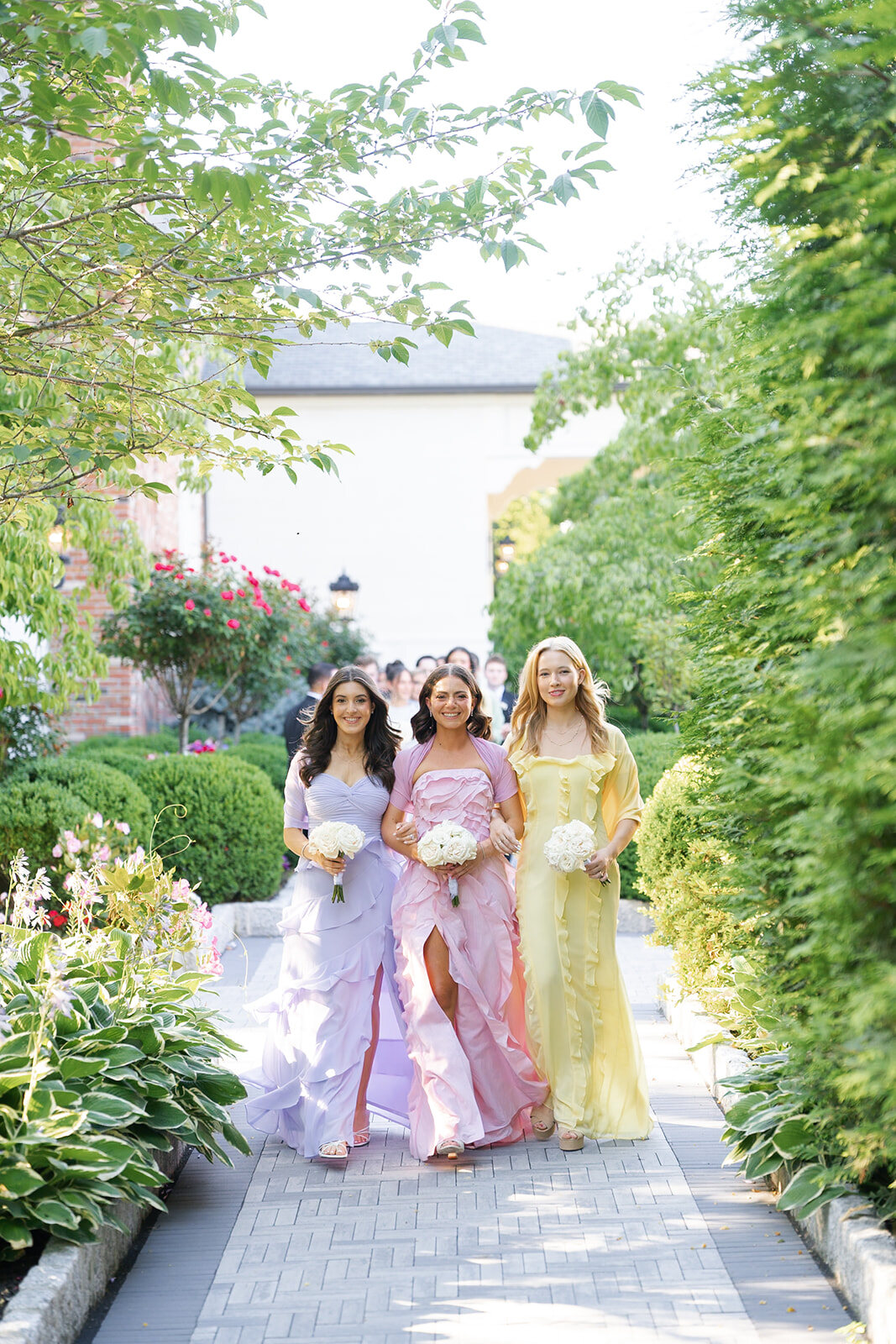 Bridesmaids in pastel gowns walk the garden aisle