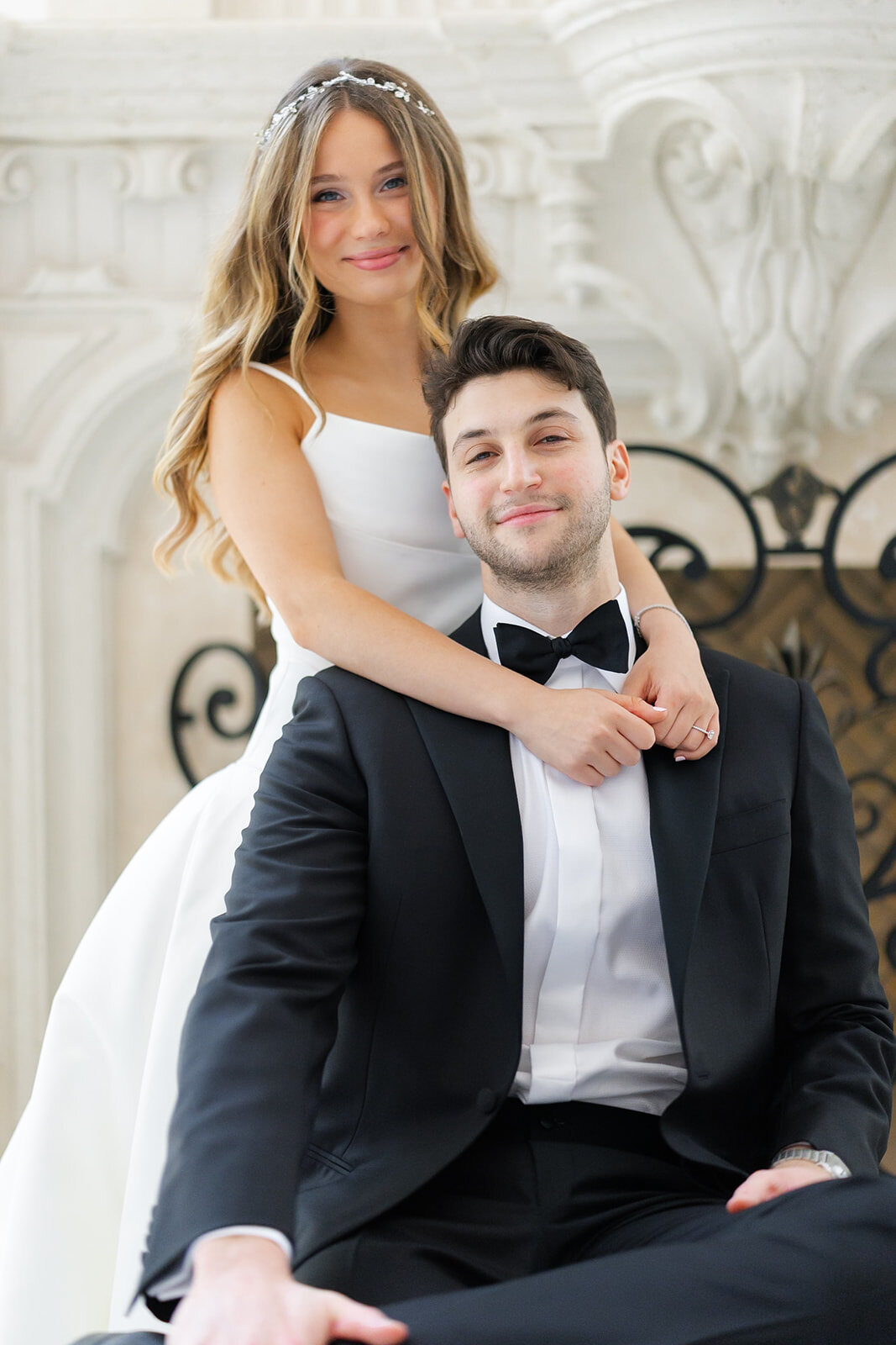 Noa leans over Nate as he sits by the carved marble fireplace