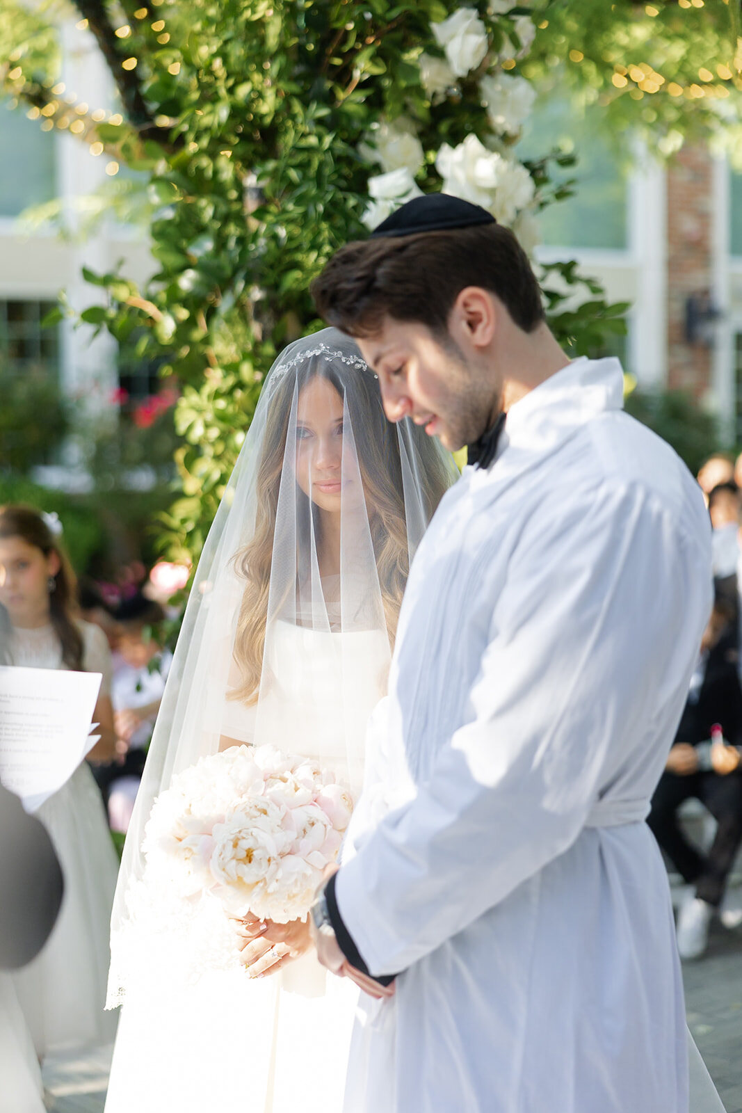 Noa, veiled, stands with Nate in his kittel under the chuppah