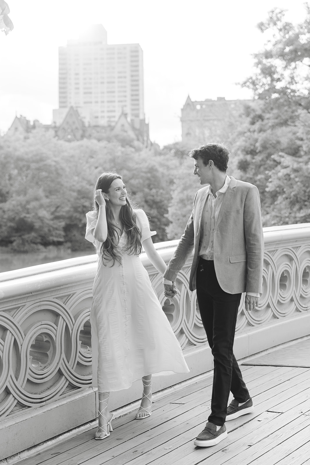 Black and white photo of an engaged couple holding hands on Bow Bridge in Central Park.