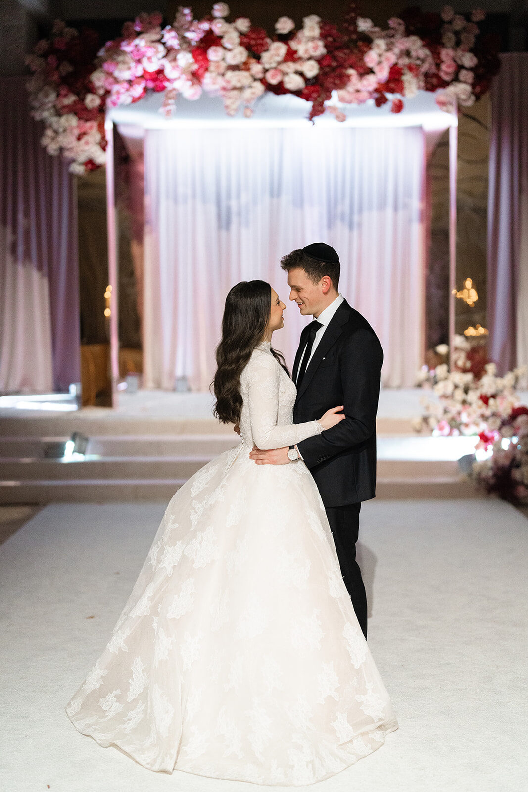Kelly and Jacob under a chuppah of pink and red roses in the ballroom at Old Westbury Hebrew Congregation