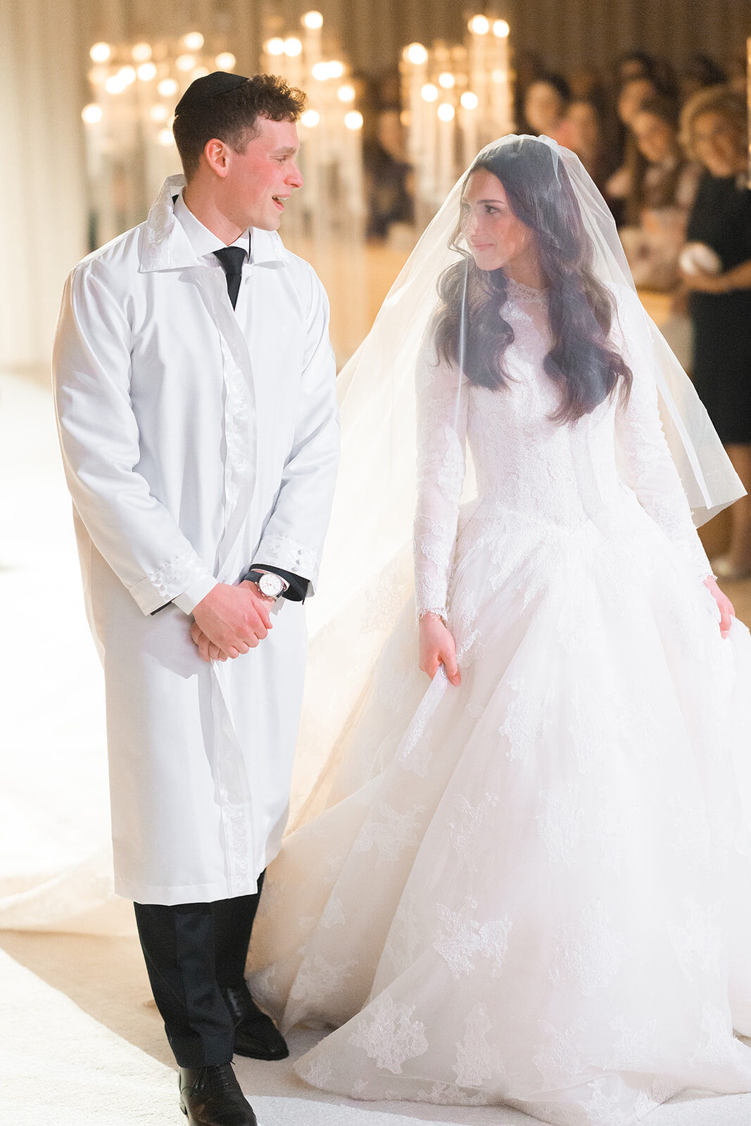 Jacob in a white kittel escorts Kelly down the aisle at Old Westbury Hebrew Congregation