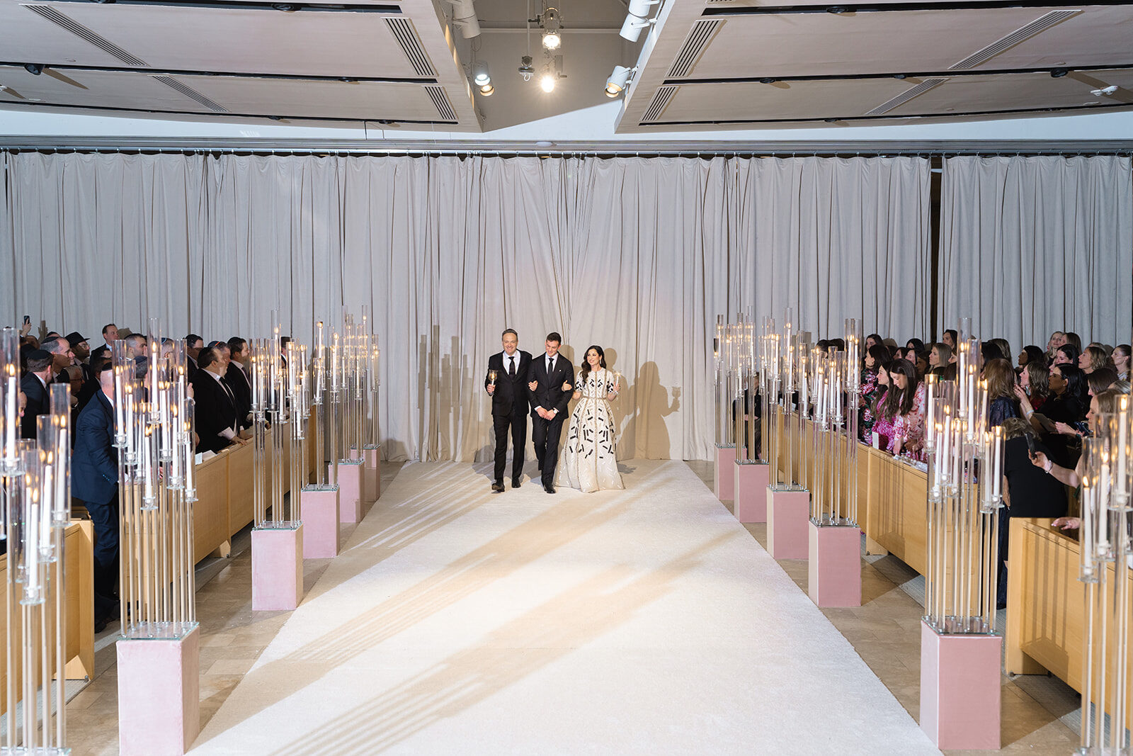 The ceremony set in the ballroom at Old Westbury Hebrew Congregation, a white aisle flanked by candles and a floral chuppah