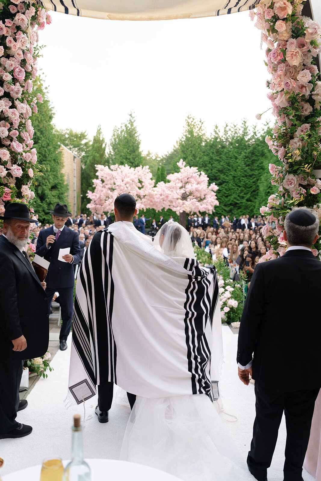 Ben in a tallit walks to the chuppah beneath pink blossoms at Old Westbury Hebrew Congregation