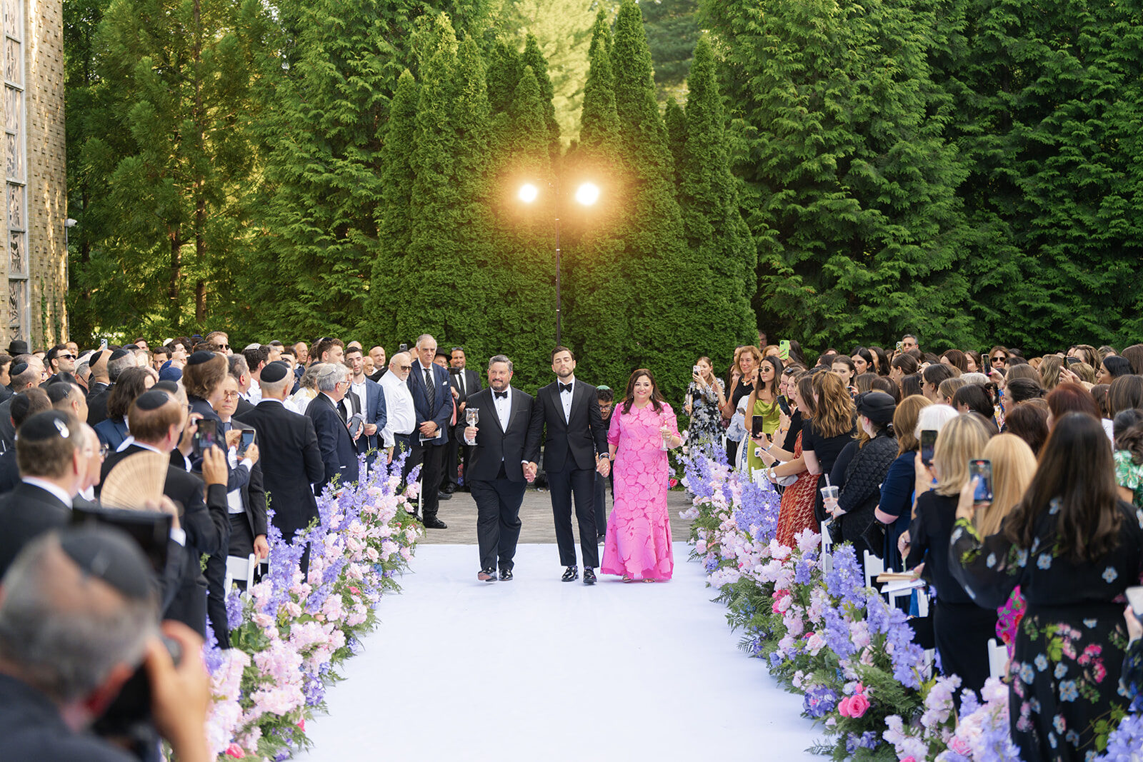 Guests seated for an outdoor ceremony at Old Westbury Hebrew Congregation, the aisle lined with purple and pink flowers