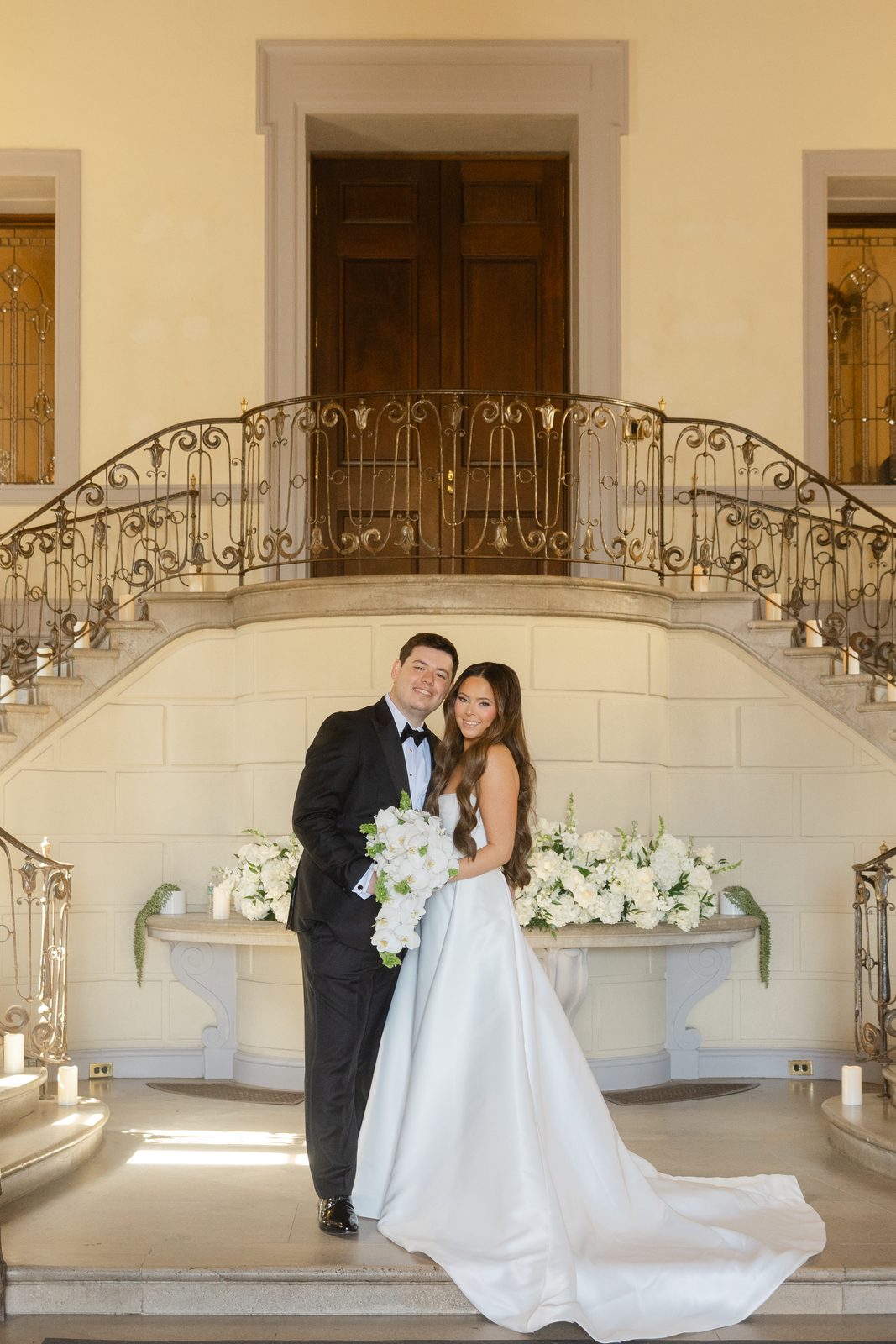 Rachel and Ian on the staircase at Oheka Castle beside white floral arrangements