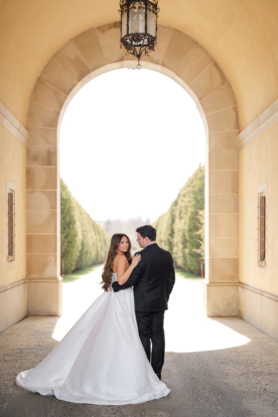 Rachel and Ian in the stone archway at Oheka Castle, the formal gardens beyond