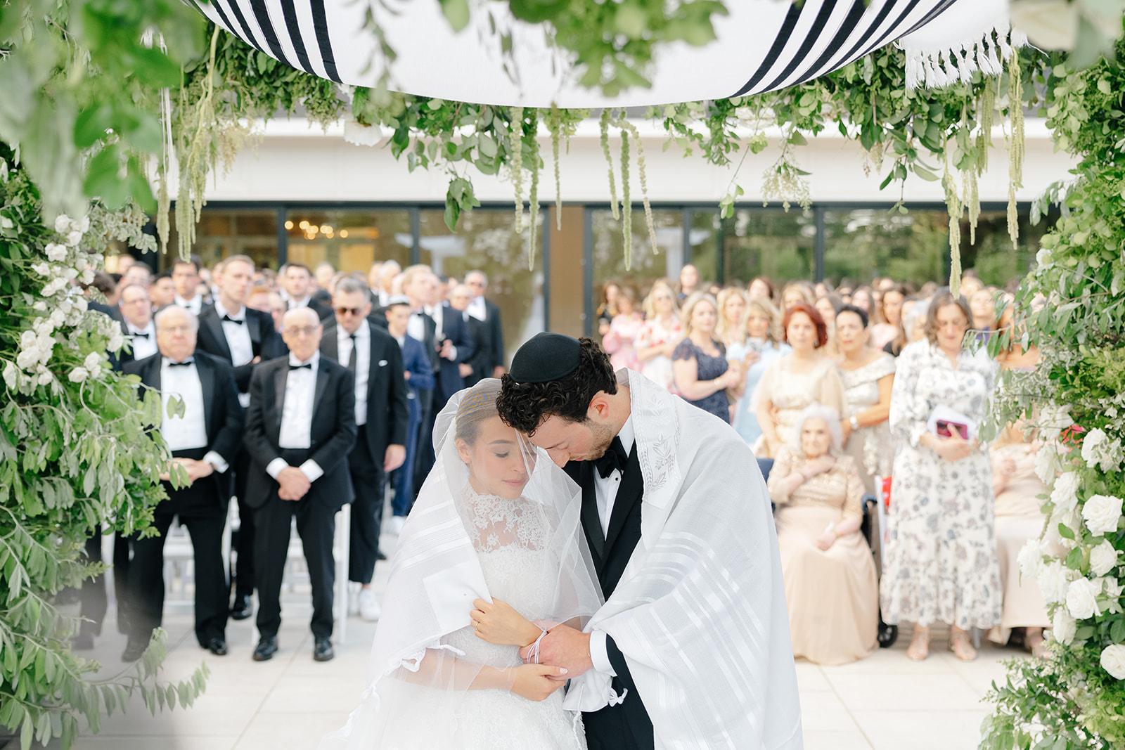 Rebecca and Phil wrapped together under the tallit-topped chuppah, surrounded by greenery and their guests