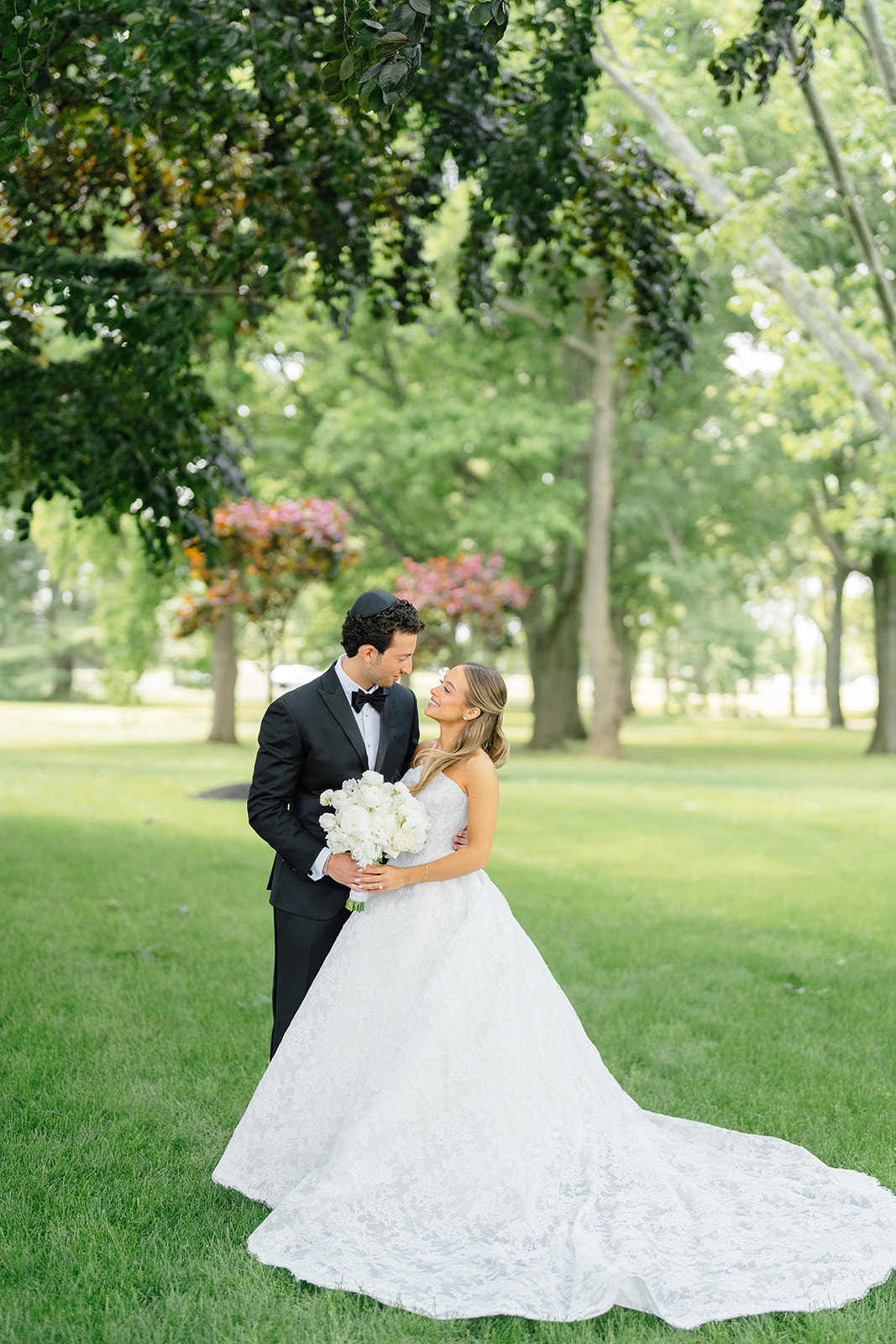Rebecca and Phil share a quiet look on a tree-lined lawn, her white peony bouquet between them