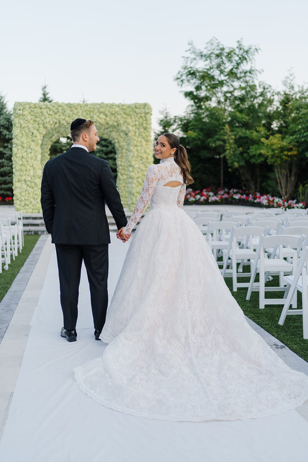 Couple walking the white ceremony aisle toward the greenery chuppah at Edgewood Country Club