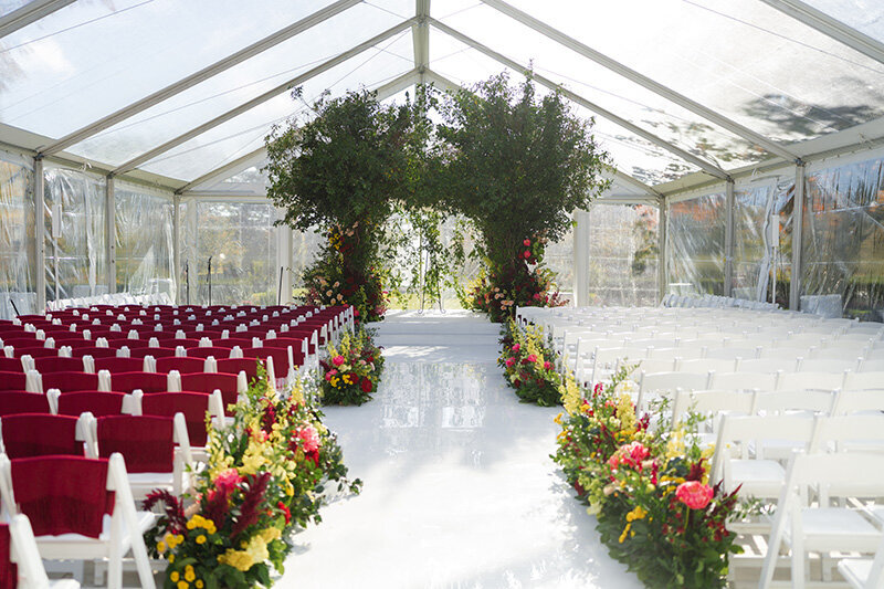 The clear-tent ceremony at The Lannin: a greenery chuppah with jewel-tone florals, a white aisle, and burgundy shawls draped on every chair.