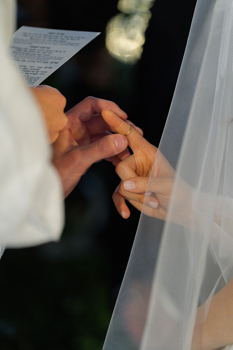 Close-up of the ring exchange under the chuppah, the bride's veil and the Hebrew ceremony program in frame.
