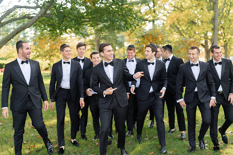 The groom walks and laughs with his groomsmen in black tuxedos under the fall trees.