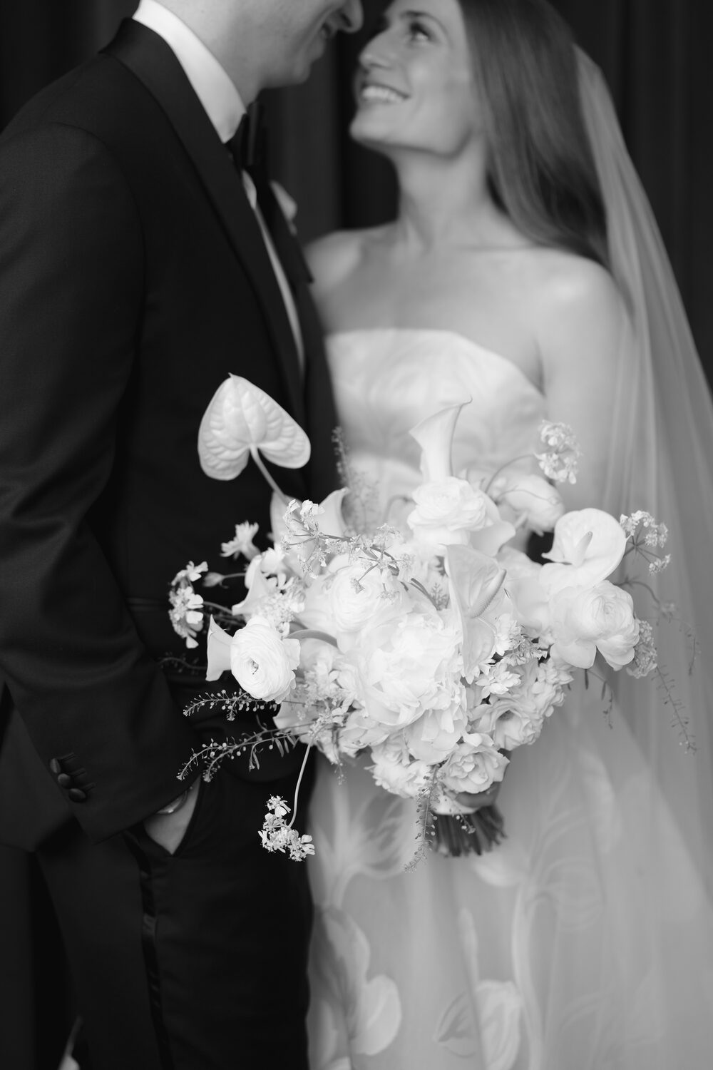 A close black and white portrait of the bride with her white bouquet, the groom just beside her.