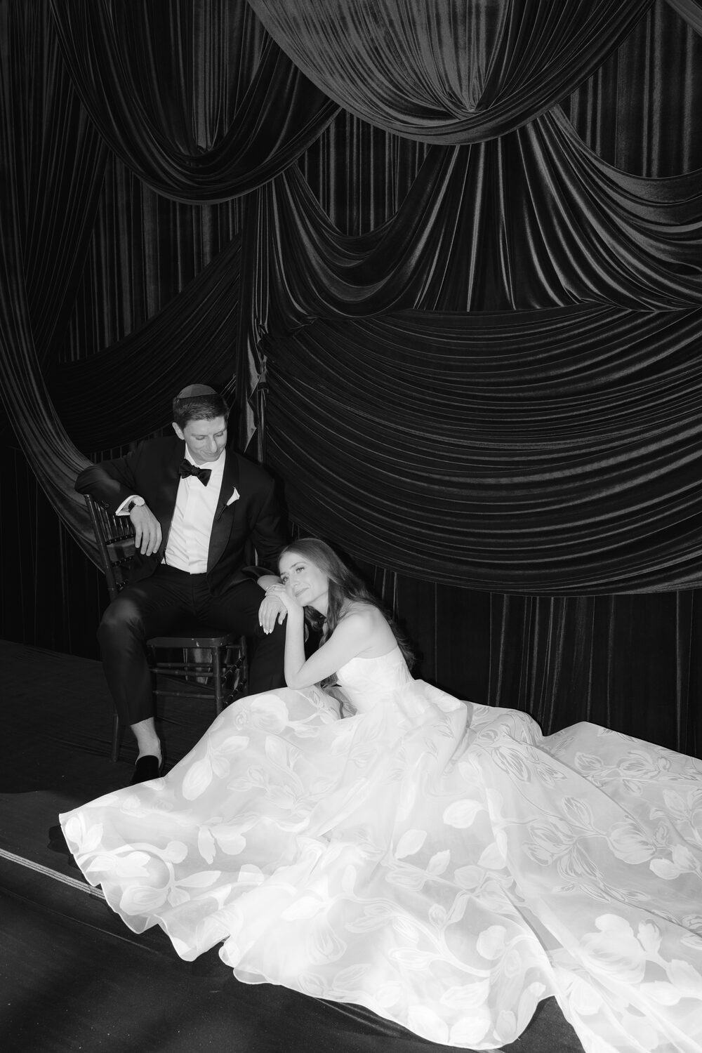 In black and white, the groom kneels to arrange the bride's train on the dark stage.