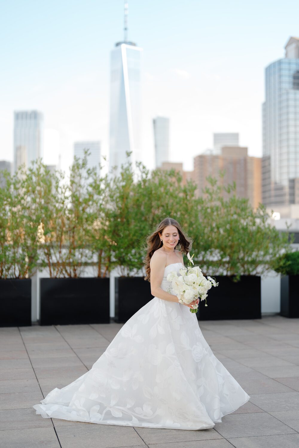 The bride holds her white bouquet on the terrace, One World Trade Center over her shoulder.