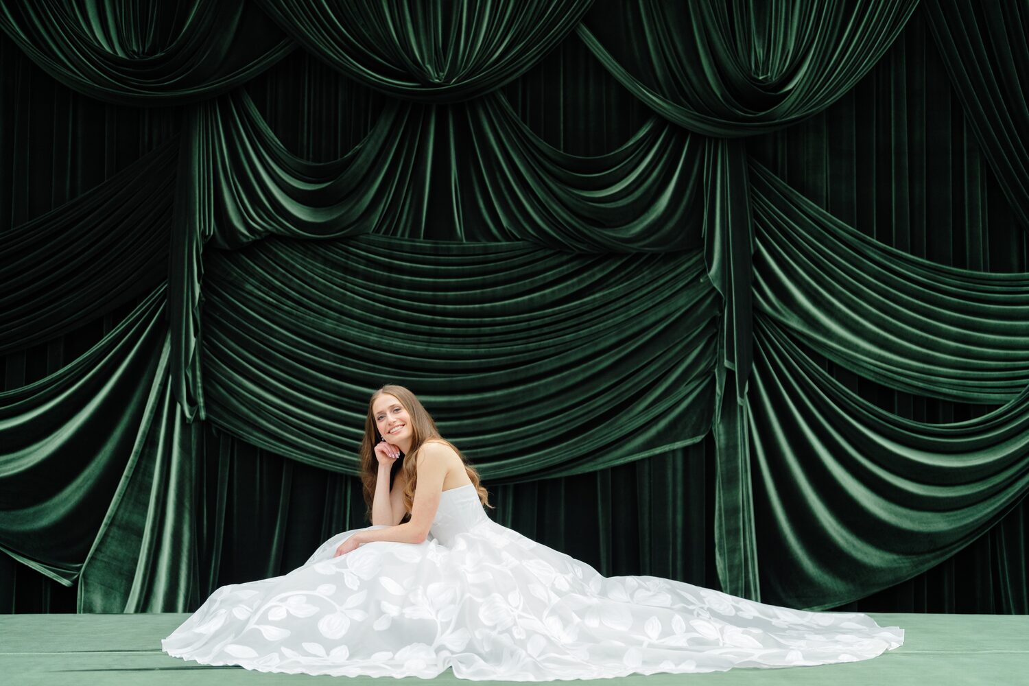 The bride sits centered on the green velvet stage, her gown spread wide around her.