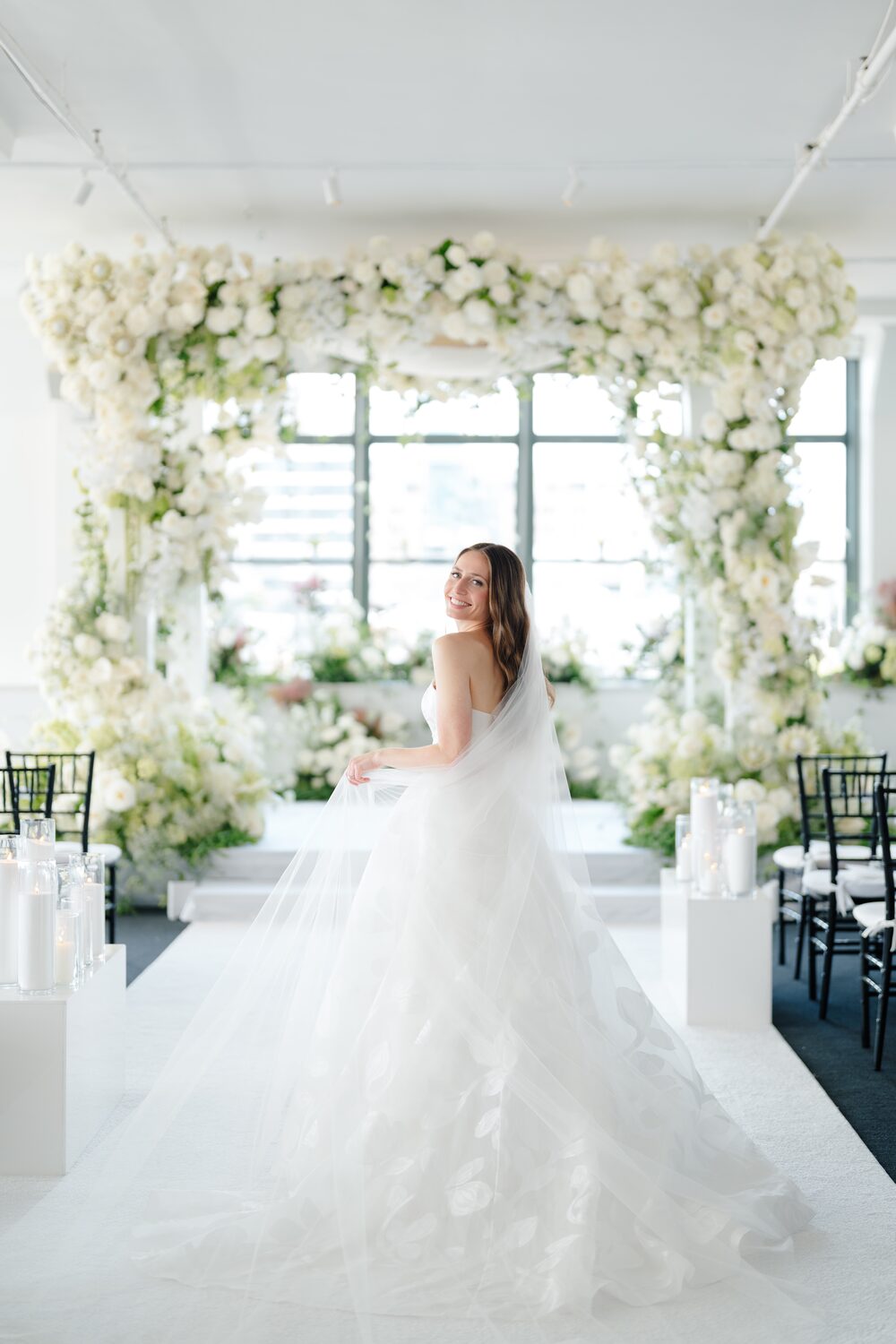 The bride glances back over her shoulder beneath the white floral chuppah.