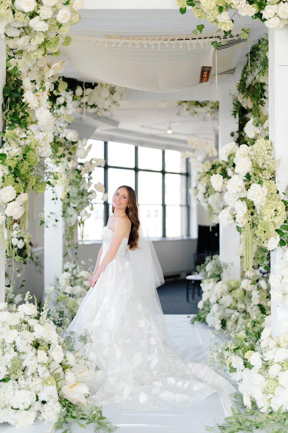 Framed by white blooms and greenery, the bride waits at the chuppah.