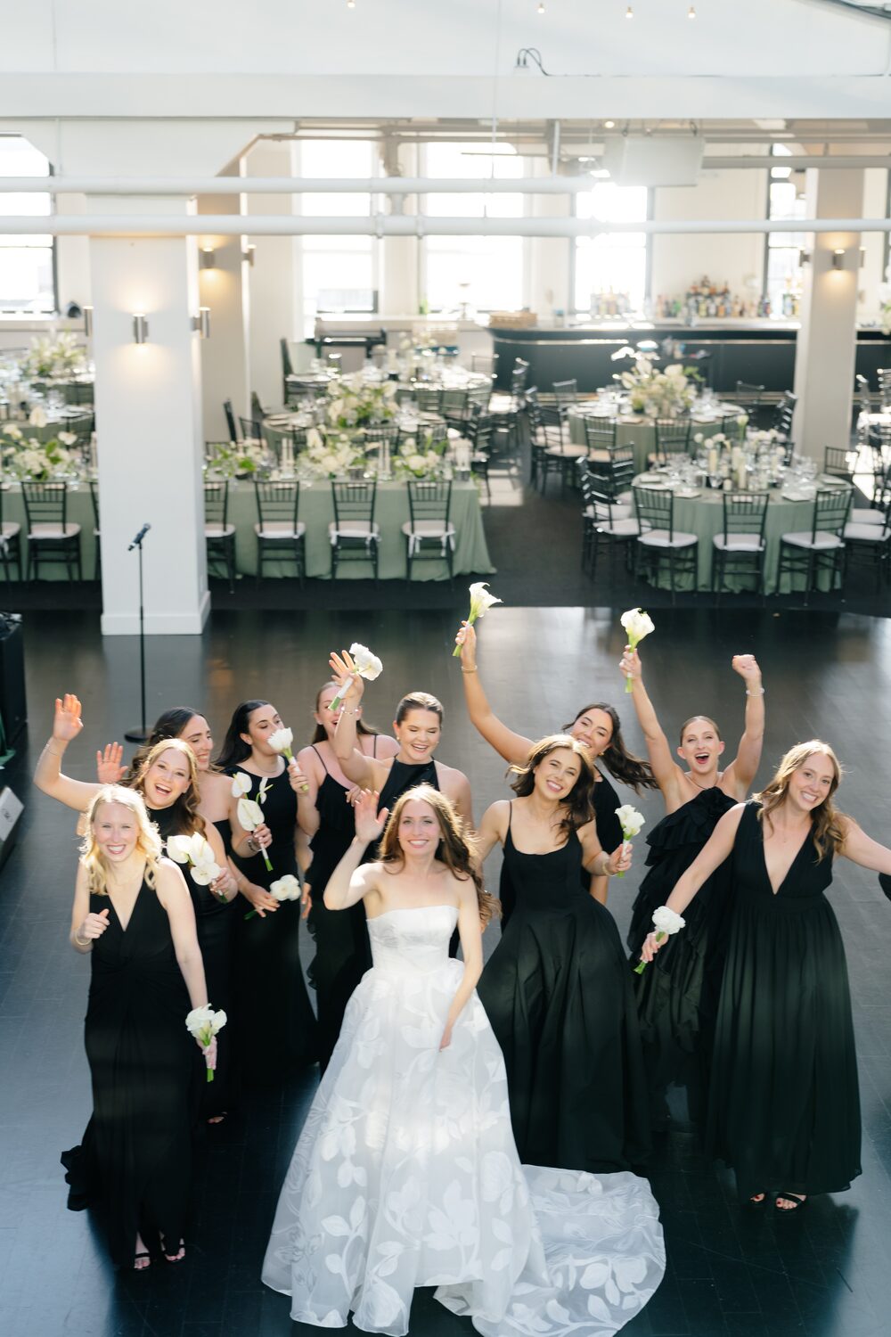 Bridesmaids in emerald gowns cheer with raised arms around the bride indoors.