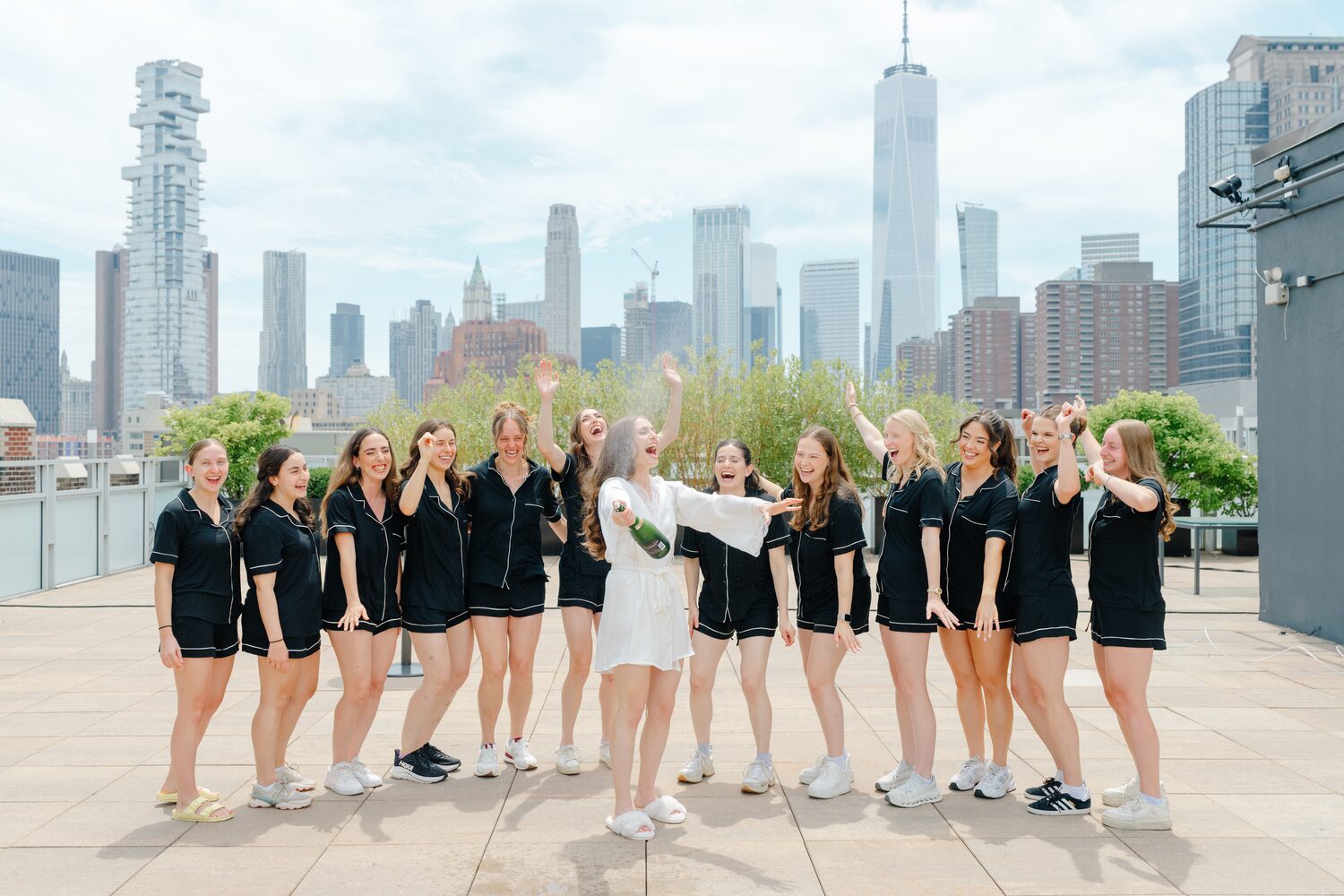 The bride and her bridesmaids in matching black loungewear line up on the Tribeca Rooftop terrace with the Manhattan skyline behind them.