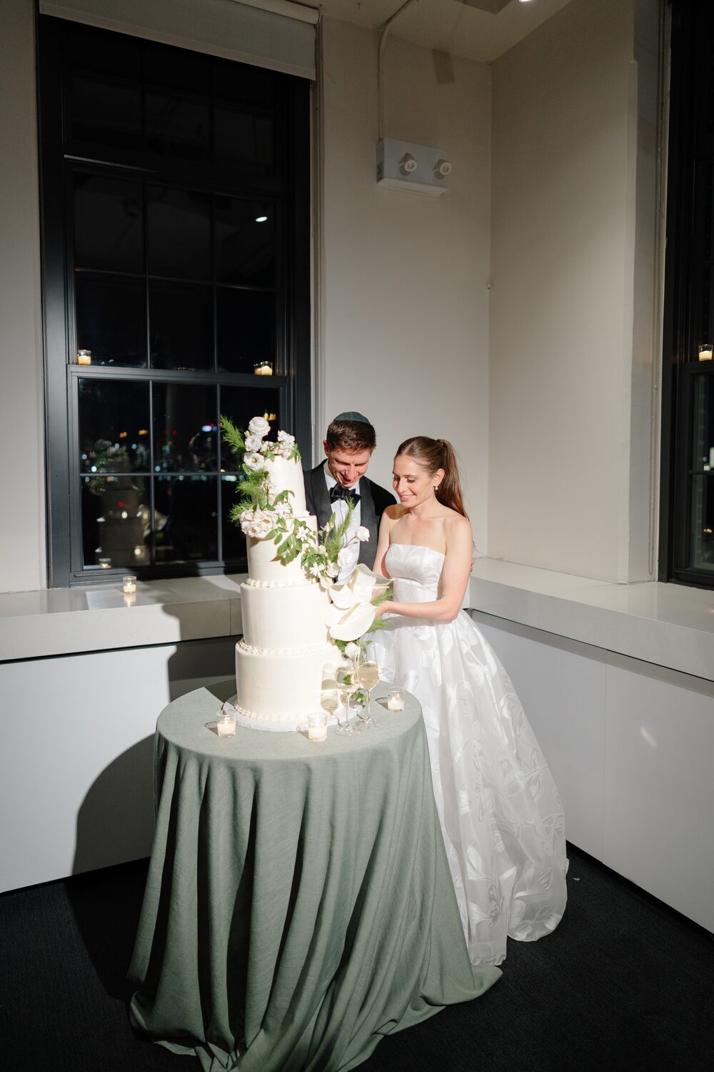 The couple cuts their cake at a sage-draped table beside the window.