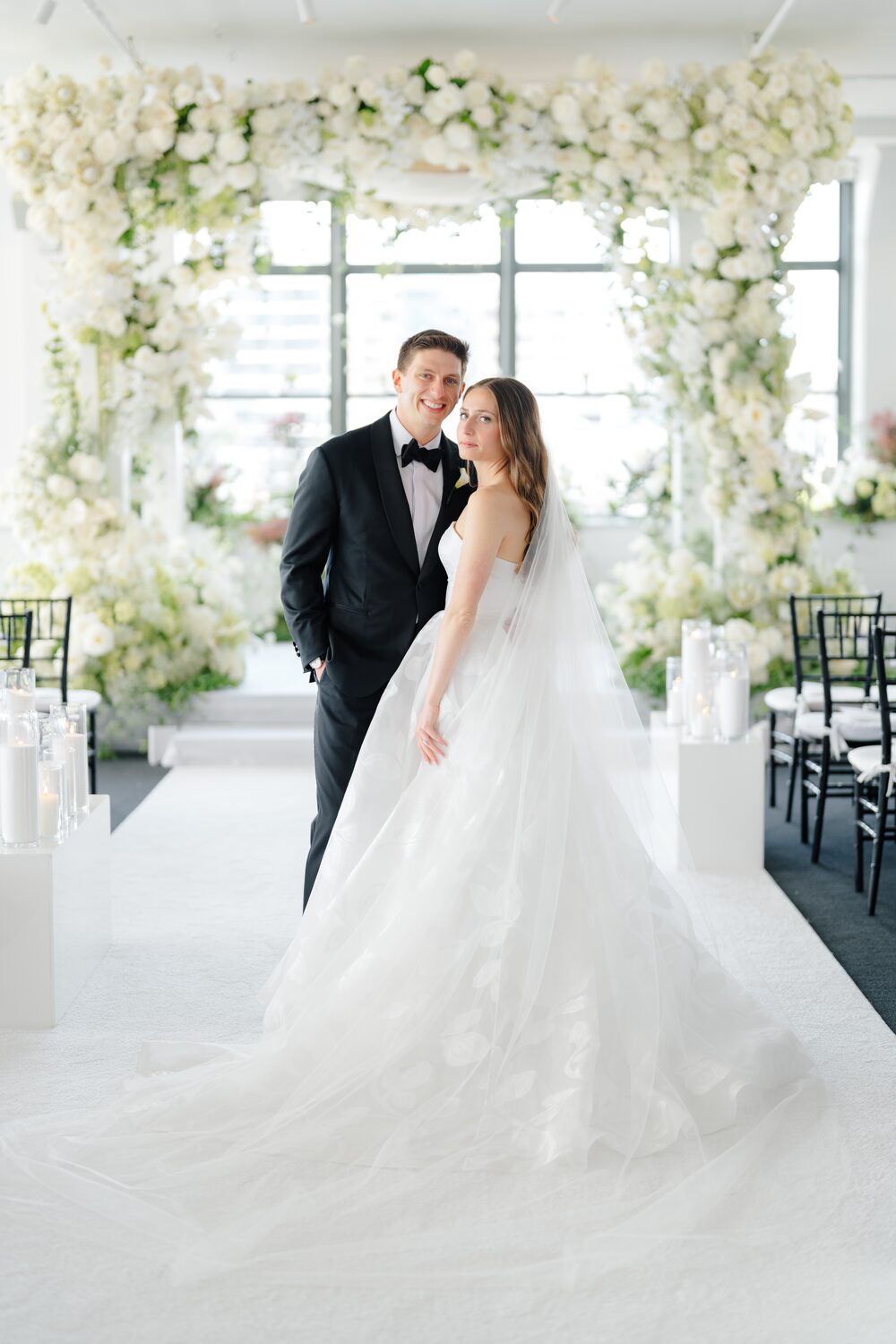 The couple stands together beneath the floral chuppah in soft window light.