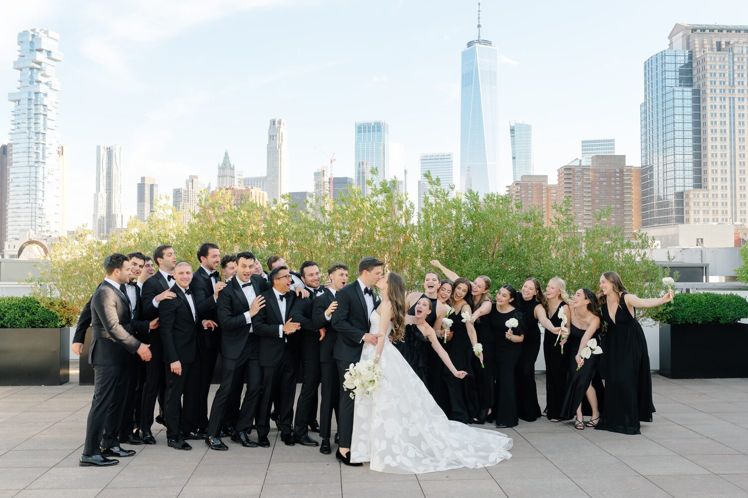 The full bridal party gathers around the couple on the rooftop with downtown Manhattan behind them.
