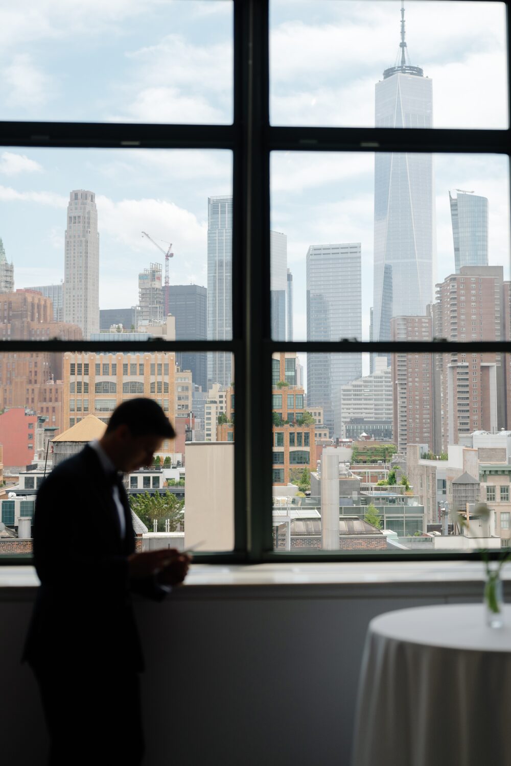The groom stands in silhouette at a tall window, One World Trade Center rising beyond the glass.