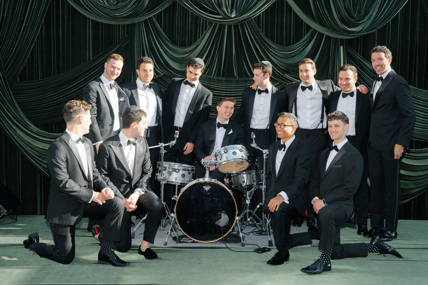 Thirteen groomsmen pose across the green velvet band stage beside the drum kit.