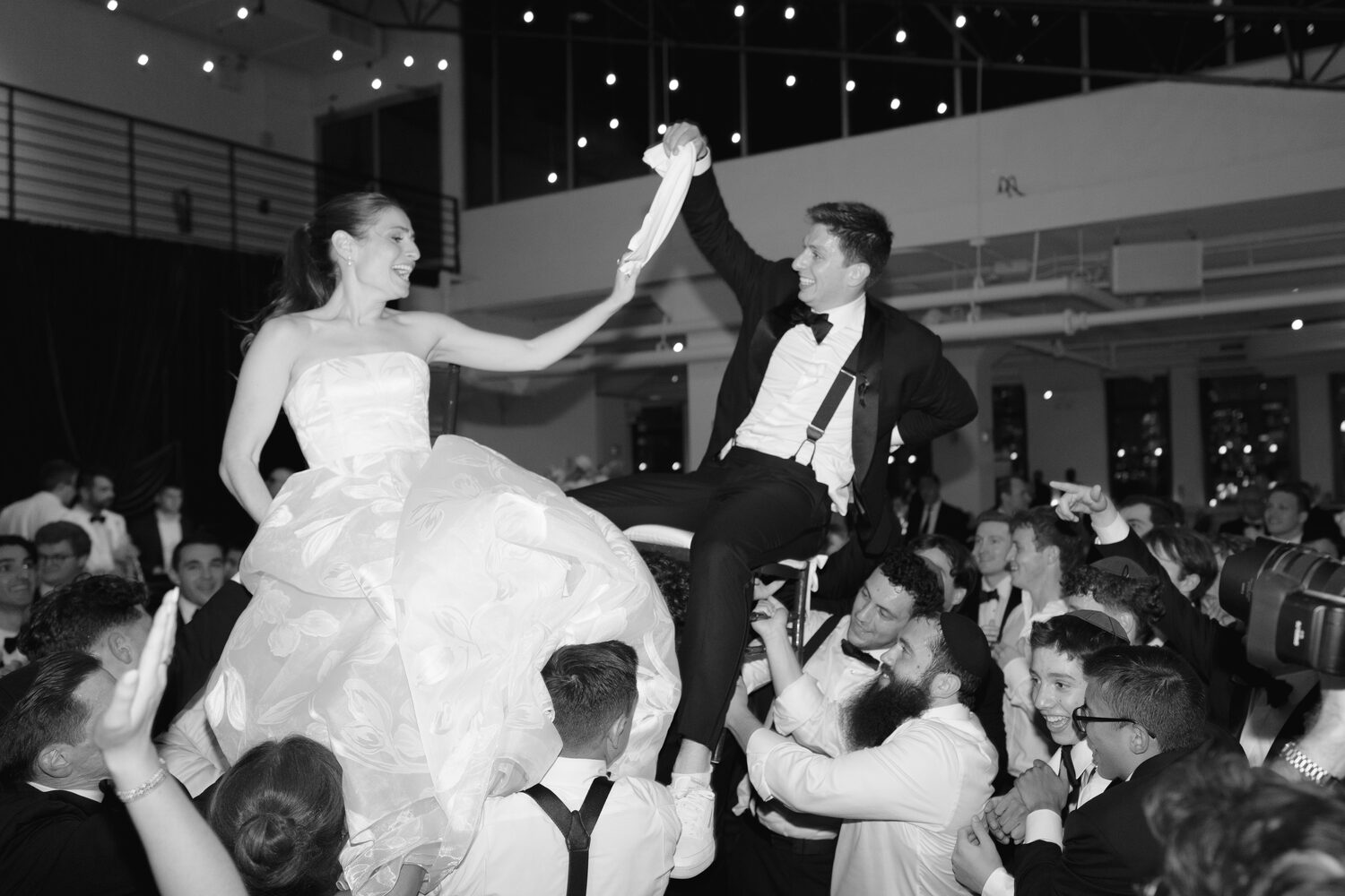 Bride and groom are lifted on chairs during the hora, hands joined above the crowd.