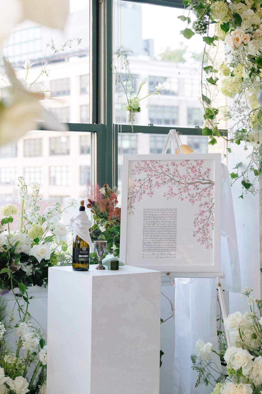 The couple's ketubah stands on display beside white florals and a skyline window.