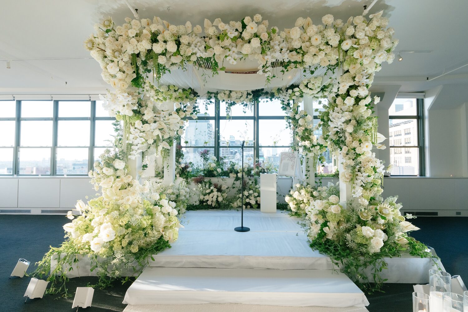 The chuppah waits under white and green florals, framed by the venue's city-facing windows.