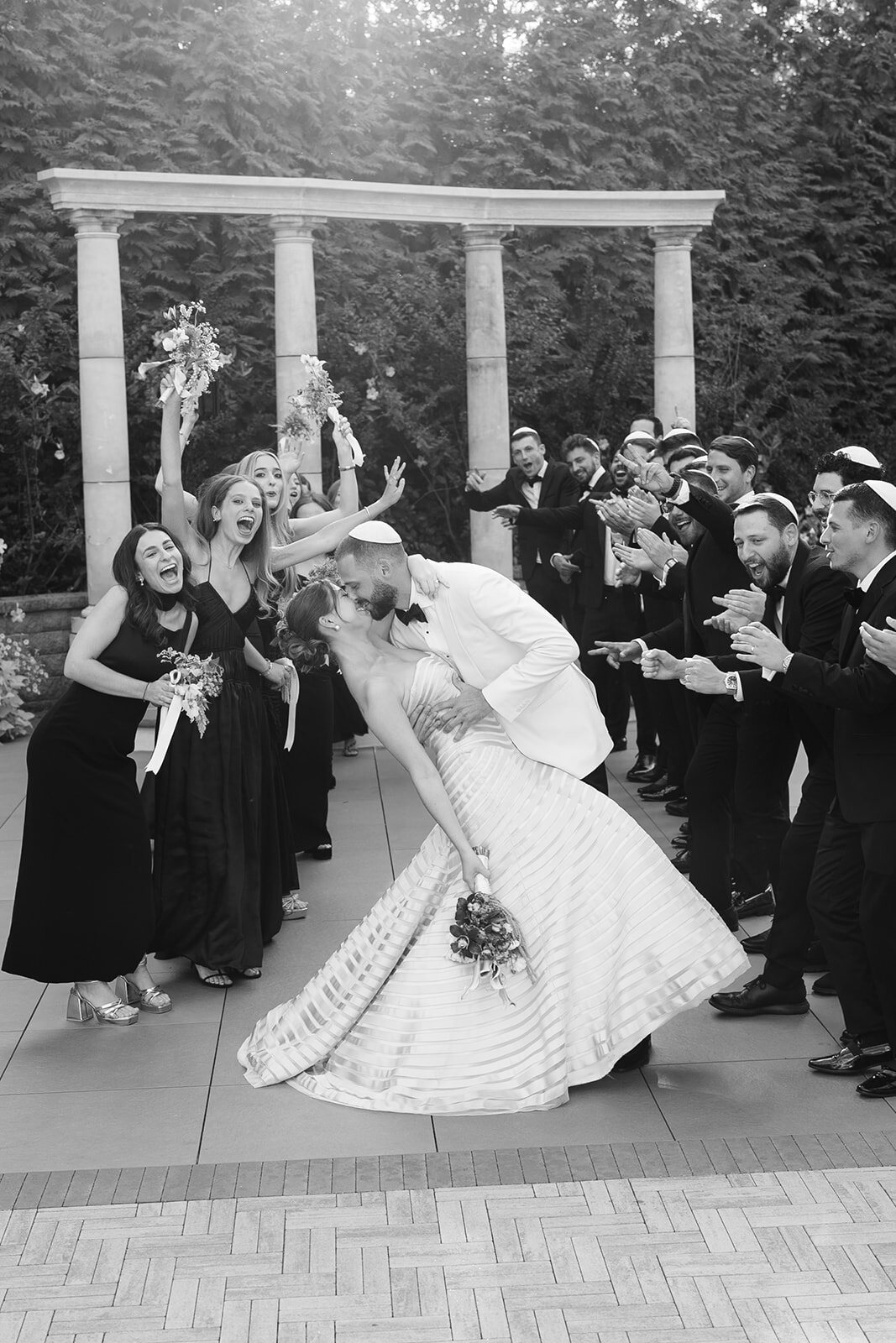Black and white photo of Yarden leaning back with her bouquet raised as the bridal party cheers