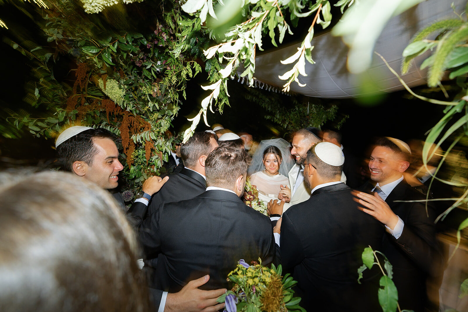 Family crowds around the couple beneath the floral canopy of the chuppah as night falls