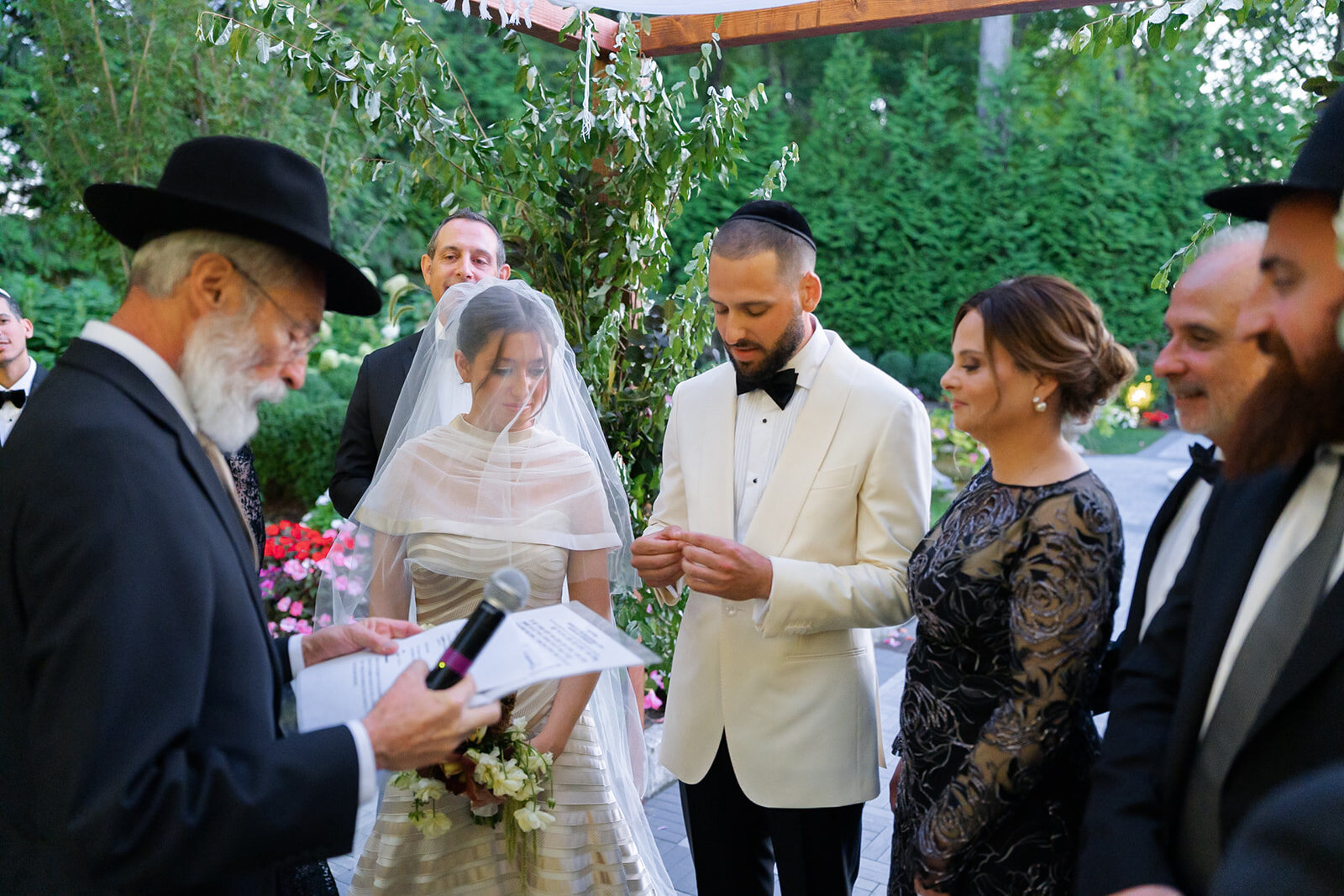 Under the chuppah, a rabbi in a black hat reads as Yarden and Joel stand together with their families