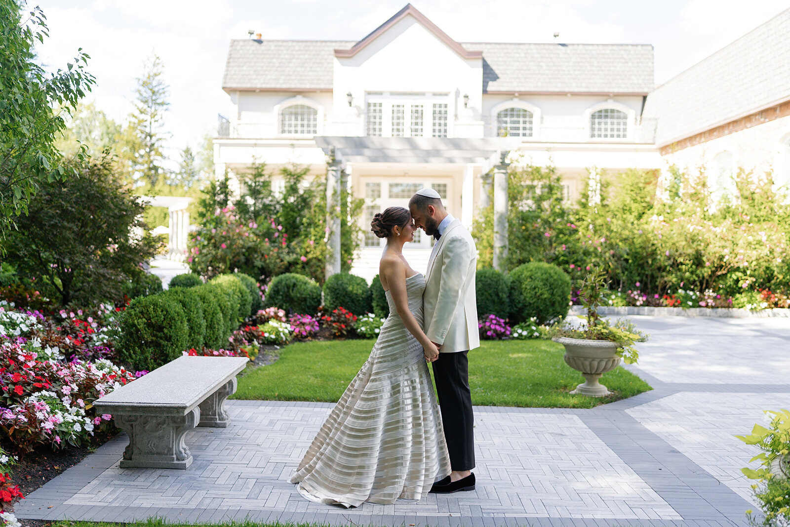 Yarden and Joel kiss on the garden path with the white Florentine Gardens manor and flower beds behind them