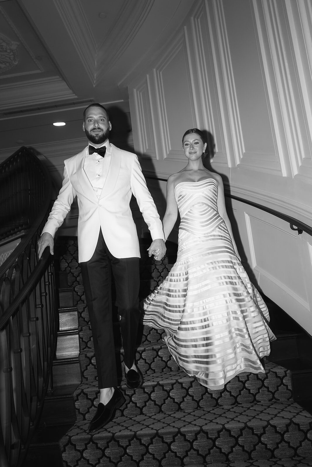 Black and white photo of Yarden and Joel descending the curved staircase hand in hand