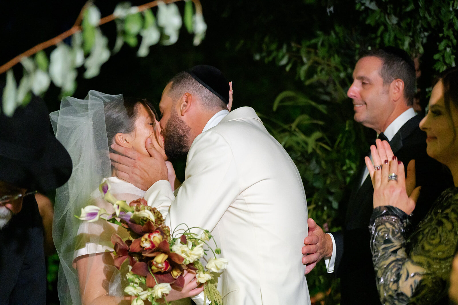 Joel kisses Yarden, her veil lifted and bouquet in hand, as guests celebrate around them