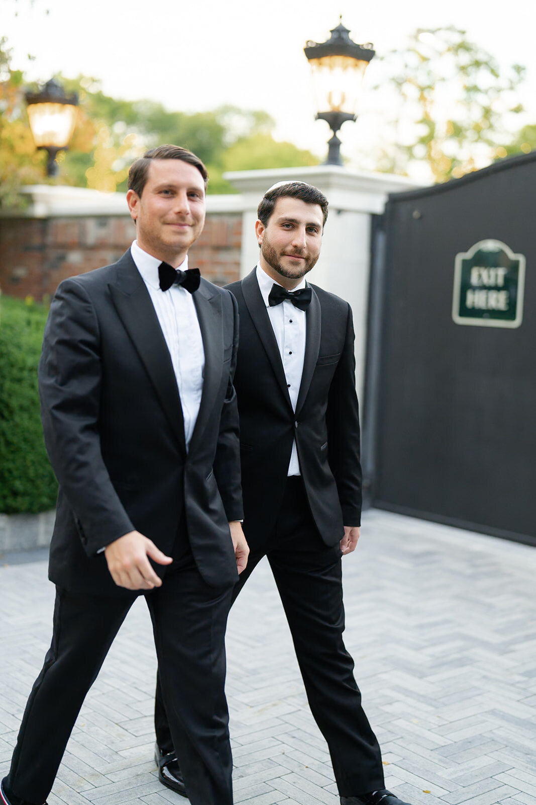 Two groomsmen in black tuxedos walk the processional past the estate gate