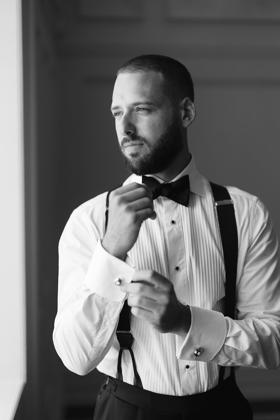 Black and white portrait of Joel fastening his bow tie in suspenders and a pleated tuxedo shirt