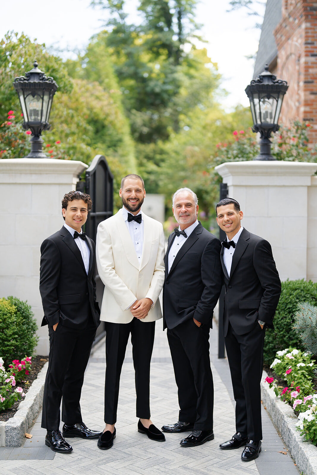 Joel in his ivory dinner jacket with his groomsmen in black tuxedos at the stone gate posts of Florentine Gardens