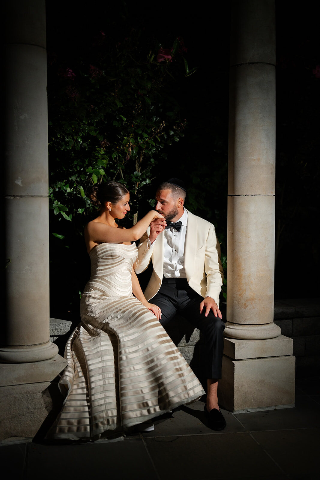 Joel kisses Yarden’s hand as she sits between the stone columns at night
