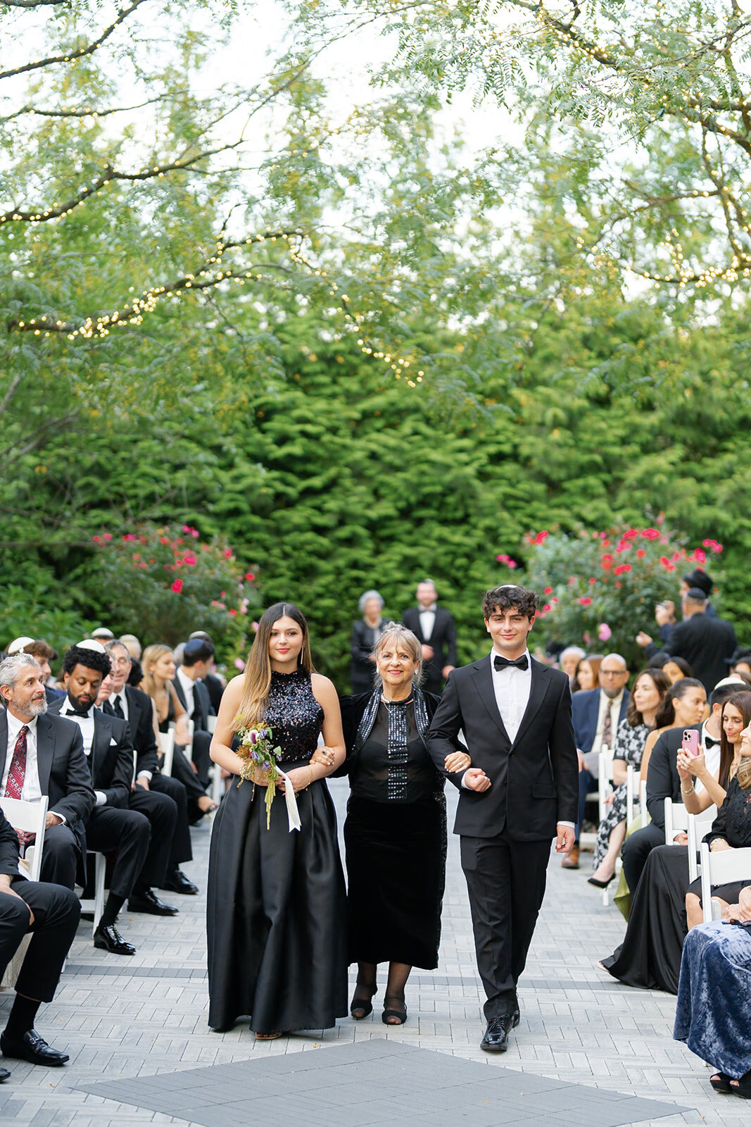 Guests line the garden aisle as the wedding party walks toward the chuppah under the trees