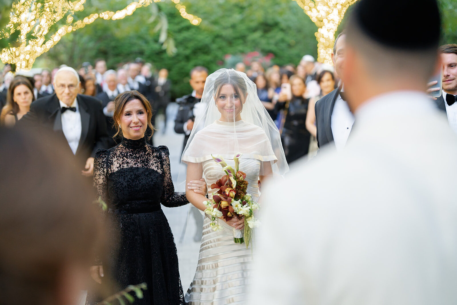 Yarden, veiled and holding her red bouquet, walks to the chuppah escorted by family
