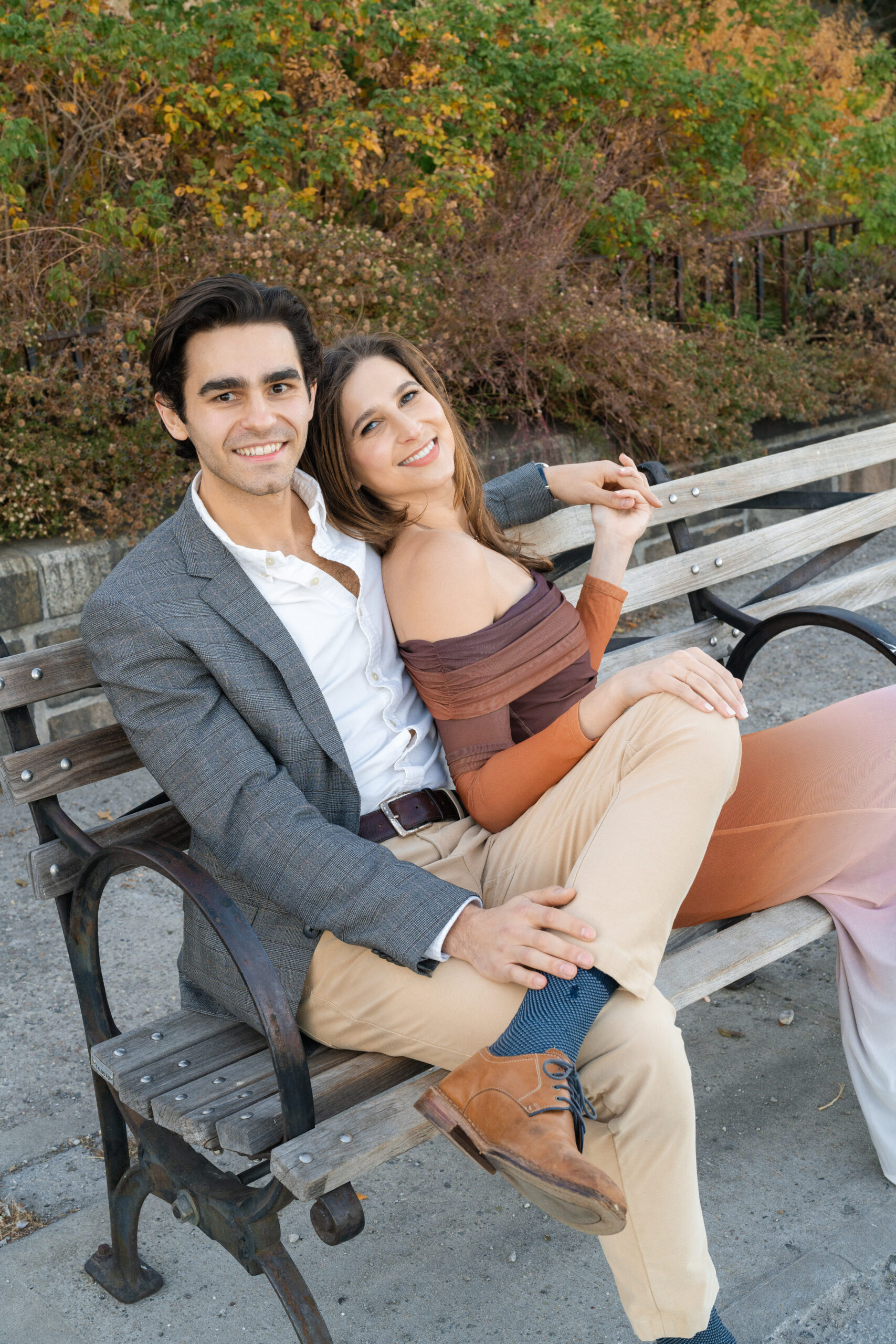 Naomi and Henry laugh together on a park bench, her in a rust ribbed top and him in a grey blazer, earth tones for a fall session