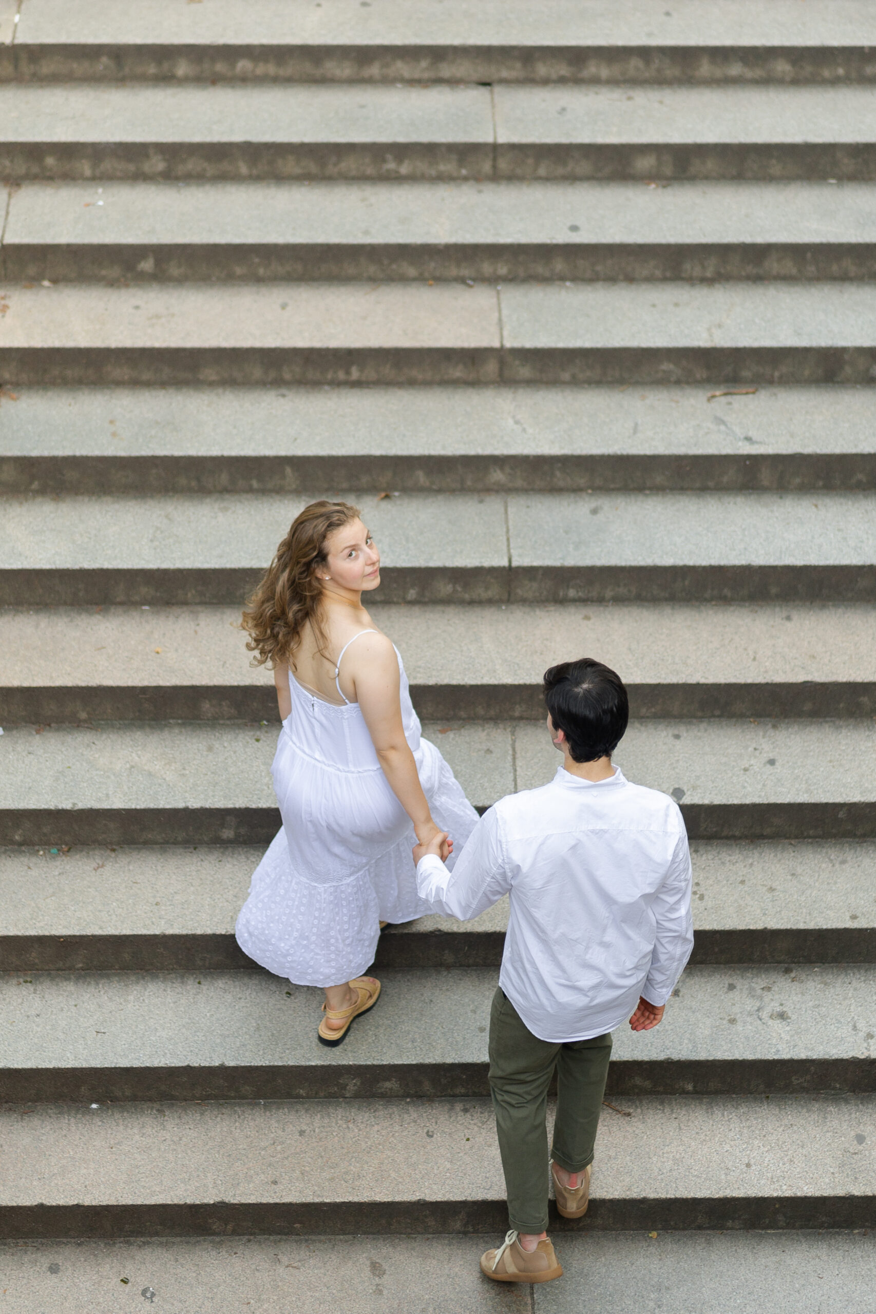 Naomi in a long pale blue dress climbs a wide stone staircase ahead of William in a white shirt, the dress moving with her