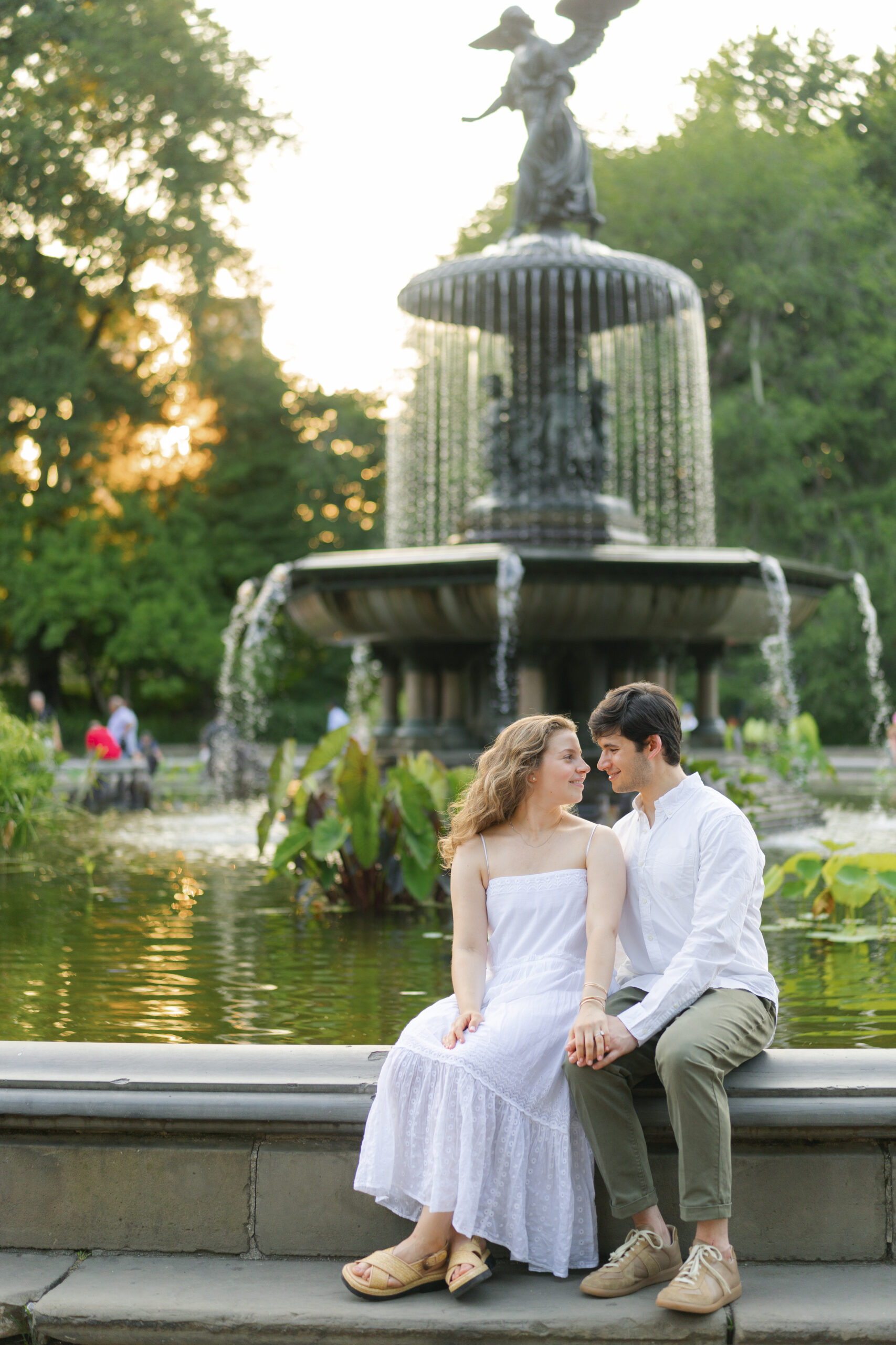 Naomi in a flowing white strapless dress seated with William at the edge of a fountain in the formal gardens, the skirt draping over the stone